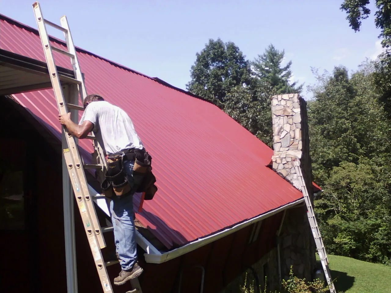 Roofer on a ladder beside a red metal roof and stone chimney — Soffit & Fascia Repair by Roofing Brothers in Lecompton